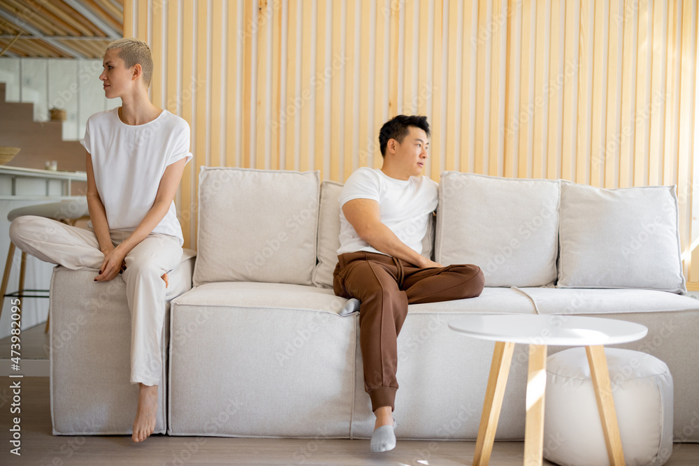 Multiracial couple sitting offended at each other on sofa at home. Concept of family relationship. Idea of domestic lifestyle. Adult asian man and young caucasian woman Multiracial couple sitting offended at each other on sofa at home. Concept of family relationship. Idea of domestic lifestyle. Adult asian man and young caucasian woman