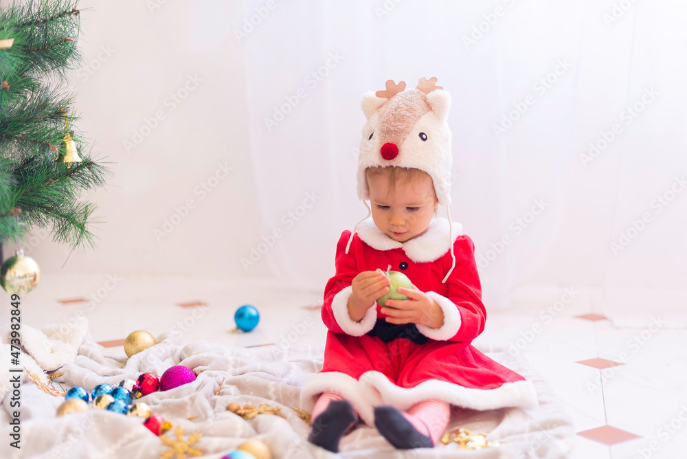 Little baby girl wearing deer hat and santa claus dress is decorating the christmas tree. 