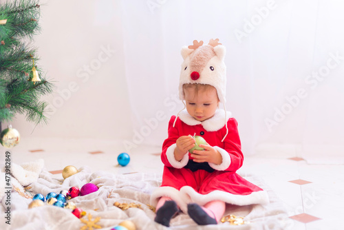 Little baby girl wearing deer hat and santa claus dress is decorating the christmas tree. 