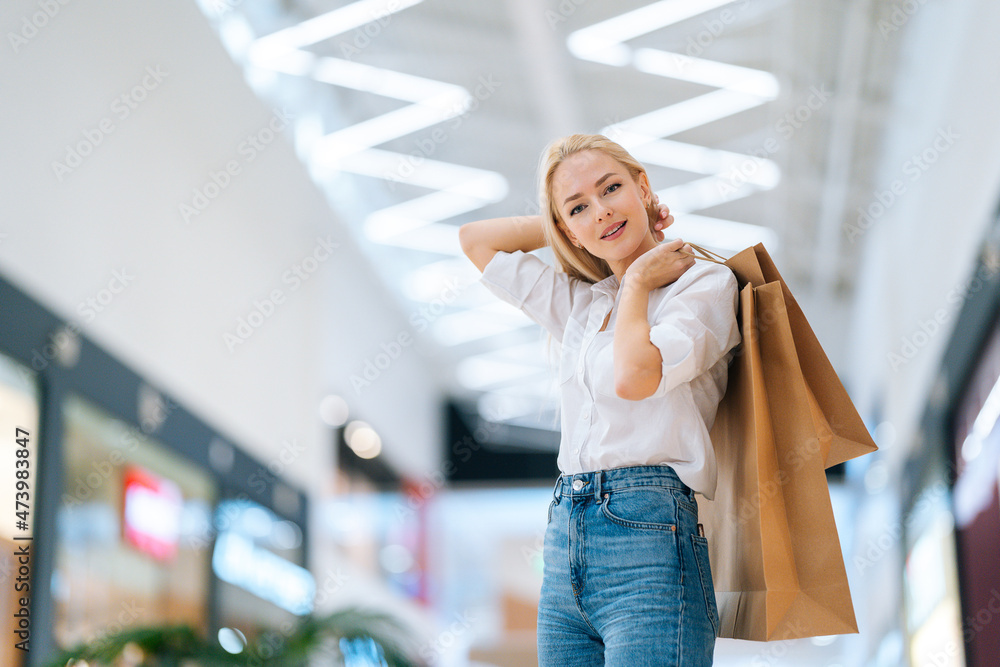 Low-angle view of attractive blonde young woman holding shopping paper bags with purchases standing in hall of mall centre, blurred background. Portrait of pretty shopaholic lady posing at store. Low-angle view of attractive blonde young woman holding shopping paper bags with purchases standing in hall of mall centre, blurred background. Portrait of pretty shopaholic lady posing at store.