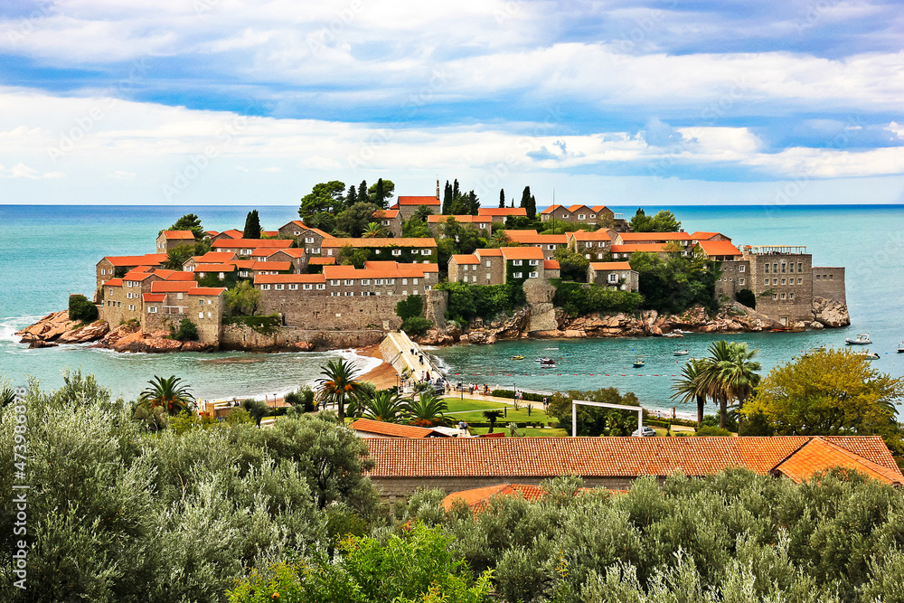 June 1, 2017. Budva, Montenegro. Picturesque view of Sveti Stefan resort village on island. View of ancient houses with red roofs against blue cloudy style. Old town architecture, green trees, nature.