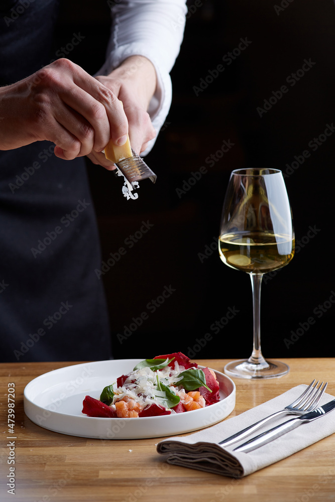 The chef prepares a salad with salmon. Rubs cheese on a grater. salmon salad with beet cheese and green leaves on wooden table with glass of white wine.