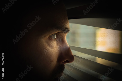 A man in a dark room looks out the window through the blinds. Covert surveillance. Close-up.