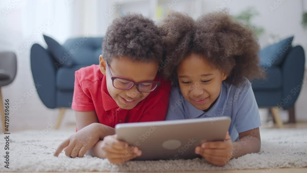 Cheerful black siblings lying on floor with tablet, having fun together, smiling Cheerful black siblings lying on floor with tablet, having fun together, smiling