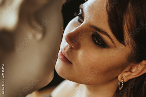Beautiful young woman kissing a plaster statue of David's head. Girl flirts with a sculpture of a bust of Apollo. Relationship with a man. Beautiful woman with bright makeup