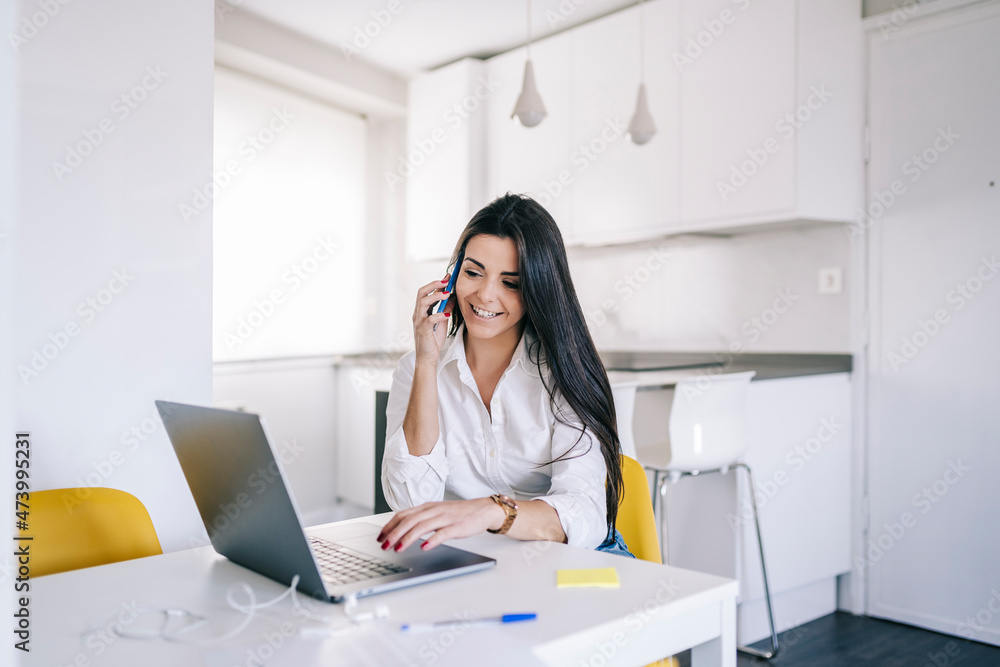 Female freelance worker with laptop talking on mobile phone at home office Female freelance worker with laptop talking on mobile phone at home office