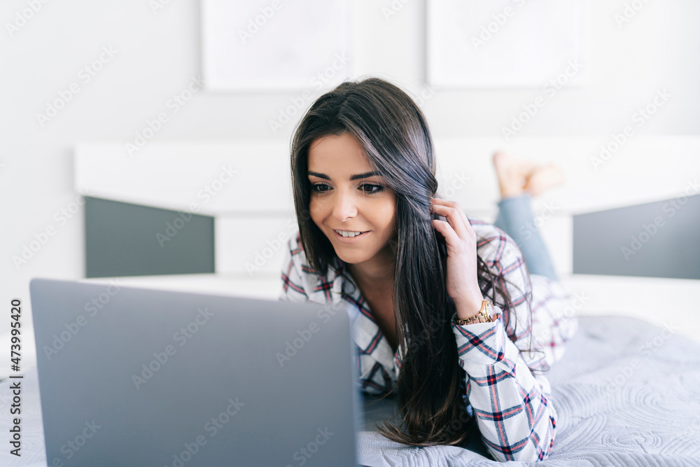 Woman with hand in hair using laptop while lying on bed at home Woman with hand in hair using laptop while lying on bed at home