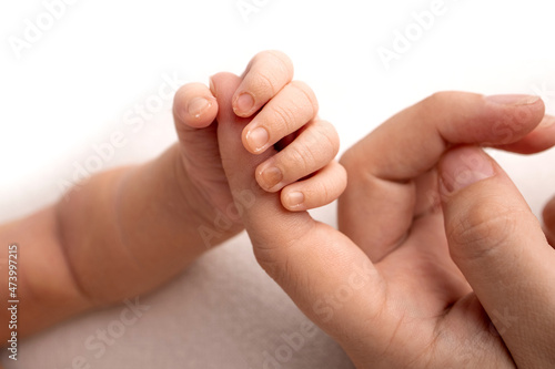 The newborn baby in a white closes has a firm grip on the parent's finger after birth. Close-up little hand of child and palm of mother and father. Parenting, childcare and healthcare concept. 