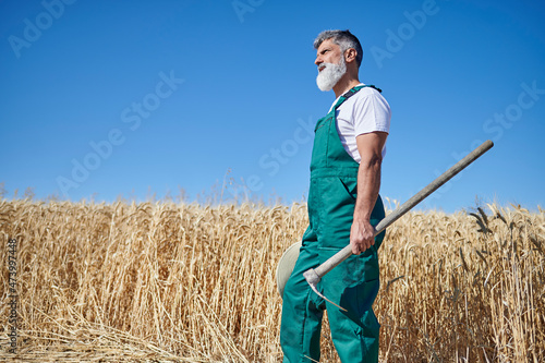 Bearded male farm worker looking away while holding hoe at wheat field during sunny day photo