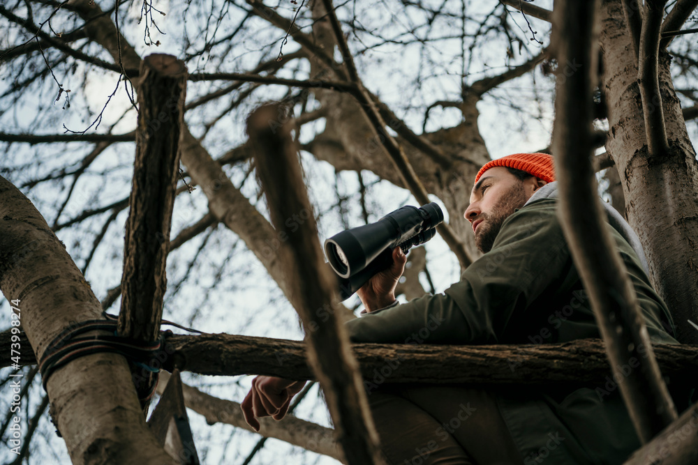 Man using binoculars while sitting on bare tree Man using binoculars while sitting on bare tree