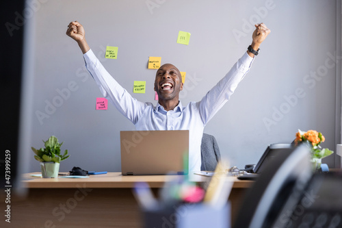 Cheerful businessman with arms raised while sitting with laptop at office