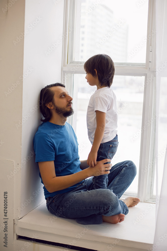 father in a blue T-shirt and a son in a white T-shirt are sitting on the windowsill large window