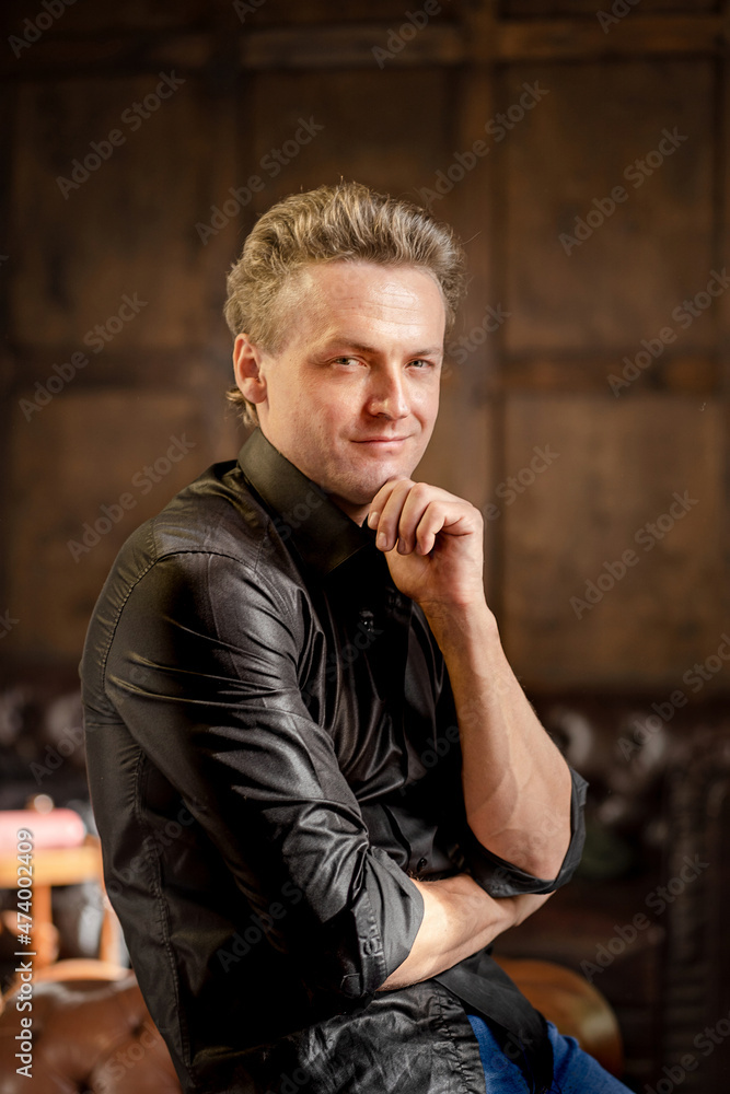 Charming Caucasian Man with Blue Eyes in a Black Shirt with Roll Sleeves Stands Half-Turn Looks at the Camera. Close-up Portrait. Wood Background