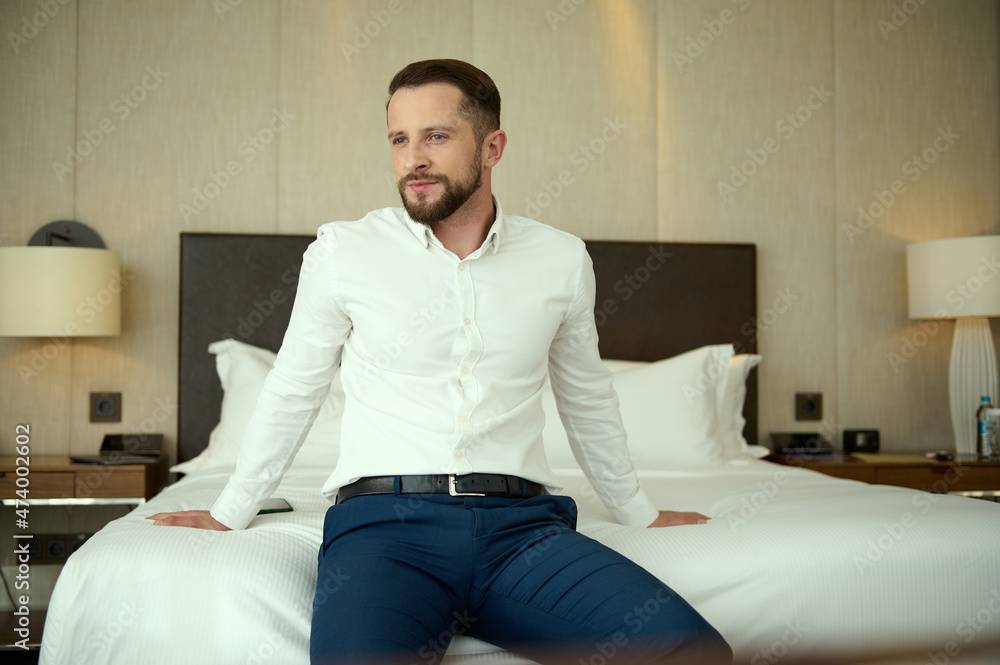Confident portrait of a handsome middle aged bearded successful businessman, entrepreneur at business trip, thoughtfully looking aside resting, sitting on the bed in hotel room