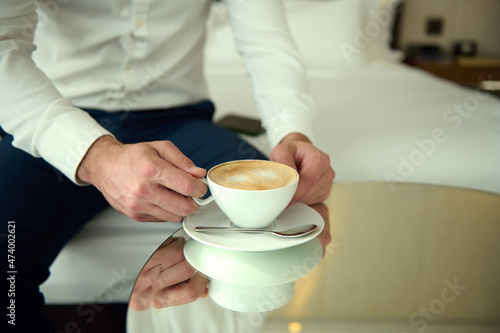 Close-up of a white ceramic cup with delicious coffee drink, cappuccino in the hands of an unrecognizable man in business casual attire, sitting on the bed in hotel room