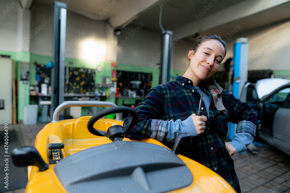 Female mechanic with screwdriver leaning on toy car at workshop Female mechanic with screwdriver leaning on toy car at workshop