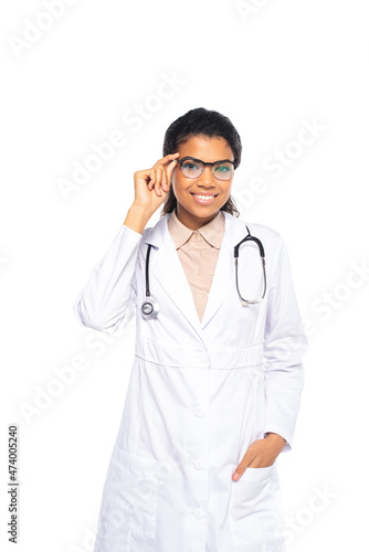 Young african american ophthalmologist holding eyeglasses and hand in pocket of white coat isolated on white.