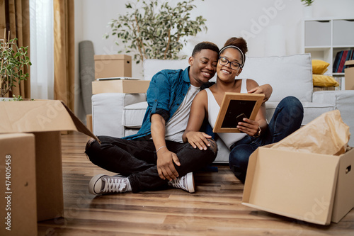 Married couple after moving to new house unpack boxes sitting on floor, woman shows husband found photo in frame from their wedding, man embraces wife, they smile reminisce