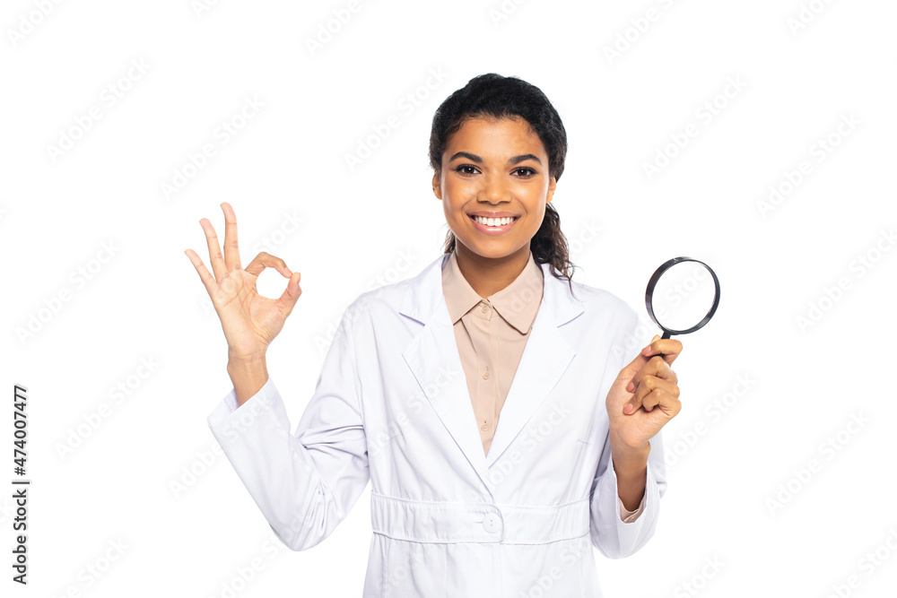 African american doctor in while coat holding magnifying glass and showing ok gesture isolated on white. African american doctor in while coat holding magnifying glass and showing ok gesture isolated on white.