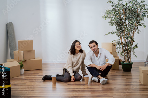 Satisfied smiling lovers sitting on wooden floor close to each other in newly purchased rented apartment drinking coffee from take-away cups getting ready to unpack moving boxes with belongings