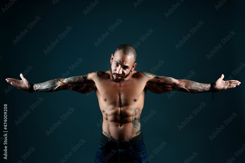 Brash muscular Latino or Cuban with tattoos on his body poses in studio against dark background. Defiant gesture and look at camera. Confident and fearless man with short haircut. Bandit.