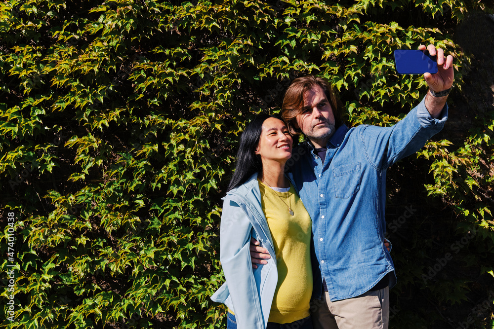 Man taking selfie with pregnant wife through smart phone in front of hedge during sunny day Man taking selfie with pregnant wife through smart phone in front of hedge during sunny day