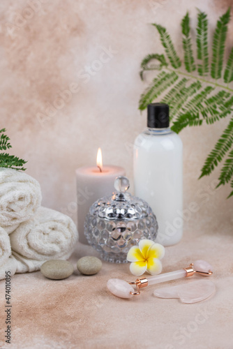 Spa still life treatment composition on massage table in wellness center. Twisted hot towel with aromatic candles and face roller gua sha on beige background. Aroma therapy setting. 