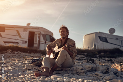 Hippy guitarist with camper house enjoying playing guitar on a sea coast in summertime.
