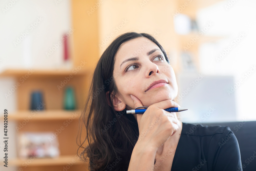 Thoughtful female professional with hand on chin looking away while sitting in office Thoughtful female professional with hand on chin looking away while sitting in office