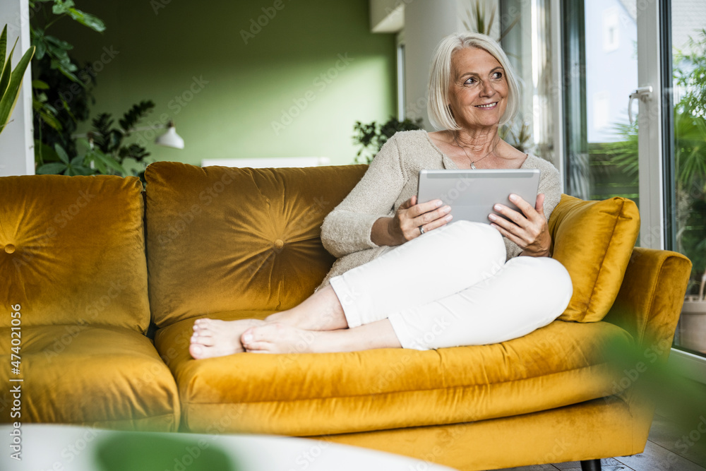 Thoughtful woman holding digital tablet while sitting on sofa in living room Thoughtful woman holding digital tablet while sitting on sofa in living room