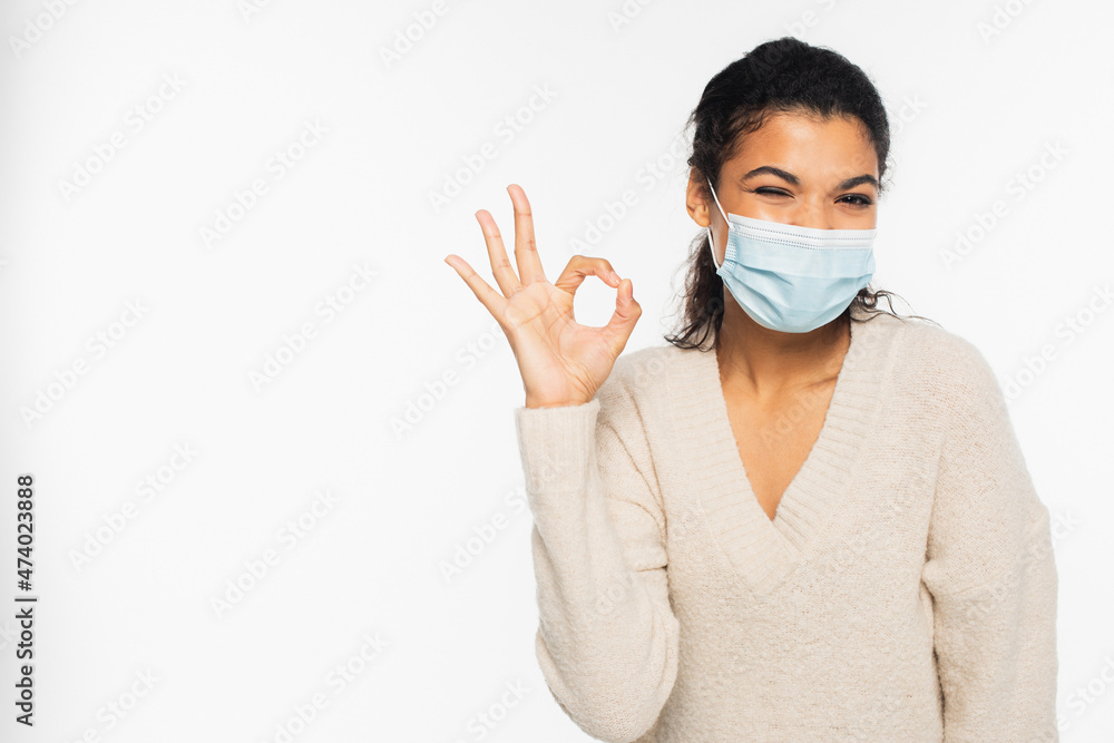 African american woman in medical mask winking and showing ok sign isolated on white.