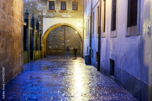 two girls walk under an arch in a narrow street in the old quarter of Oviedo one night after the rain
