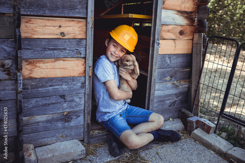 Cute boy in yellow hardhat holding rabbit in wooden hutch photo