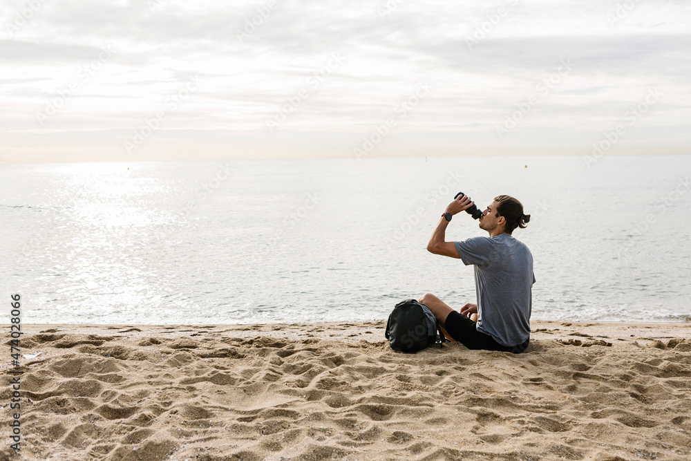 Young sportsman drinking water while sitting on beach Young sportsman drinking water while sitting on beach