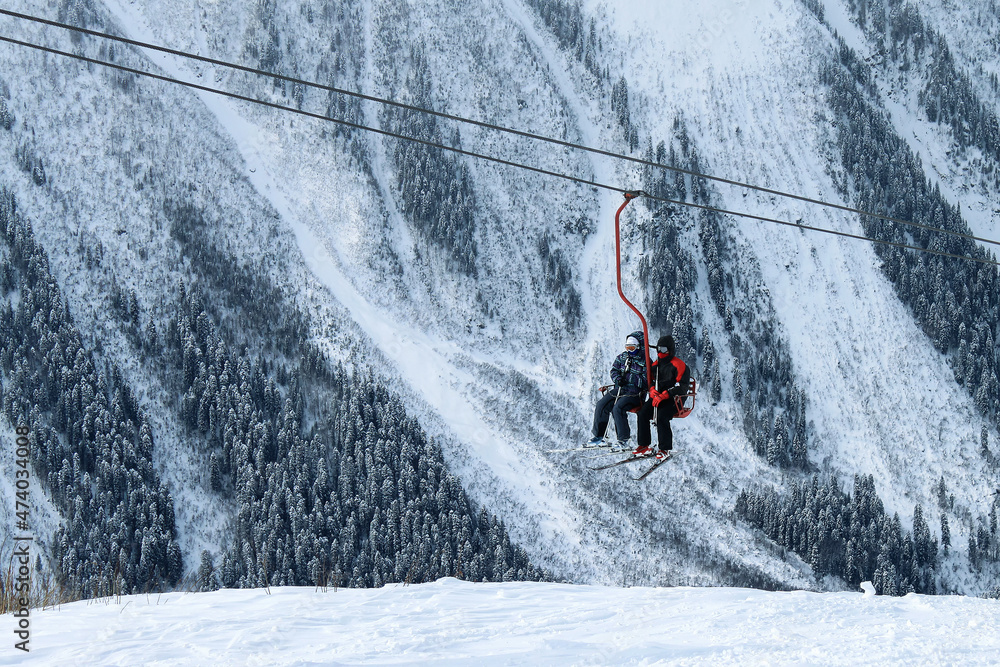 Two skiers ride a chairlift in front of a mountain with snow-covered fir trees. Winter recreation at a ski resort.