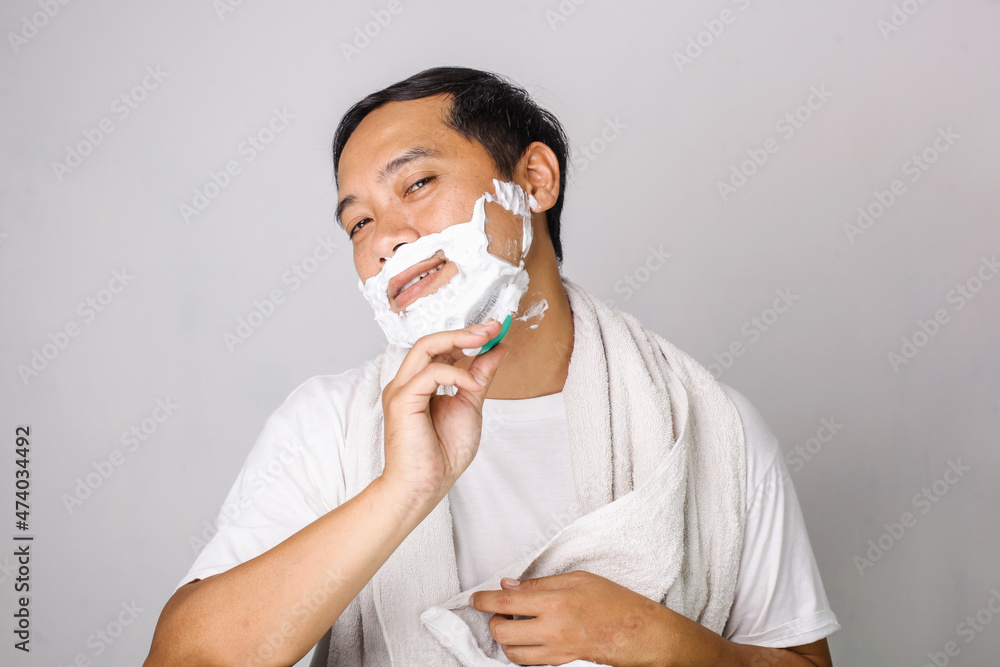 Handsome asian man shaving. Handsome asian man shaving his face and looking at camera while standing on grey background Handsome asian man shaving. Handsome asian man shaving his face and looking at camera while standing on grey background