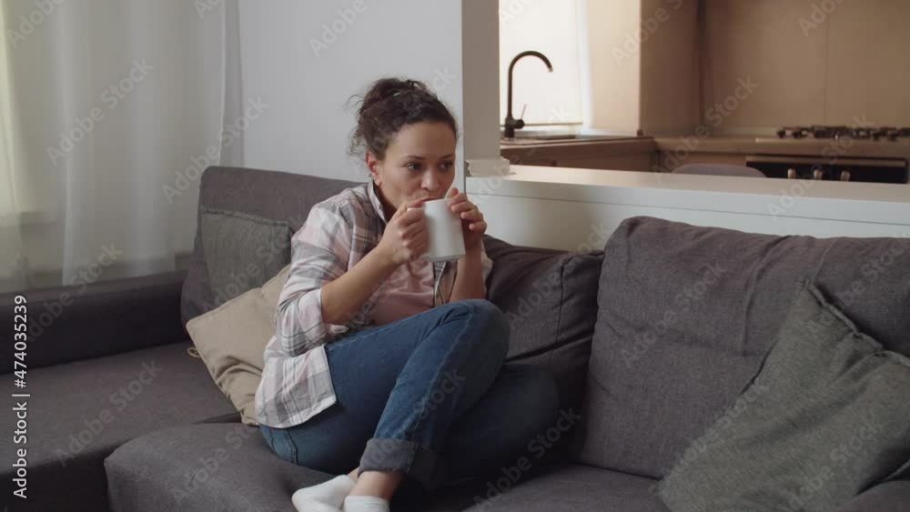 Adult attractive woman holding white mug, drinking hot beverage, sitting on couch, enjoying pastime at home. Good looking lady in casual clothing savoring cup of hot coffee or tea, daydreaming indoors Adult attractive woman holding white mug, drinking hot beverage, sitting on couch, enjoying pastime at home. Good looking lady in casual clothing savoring cup of hot coffee or tea, daydreaming indoors