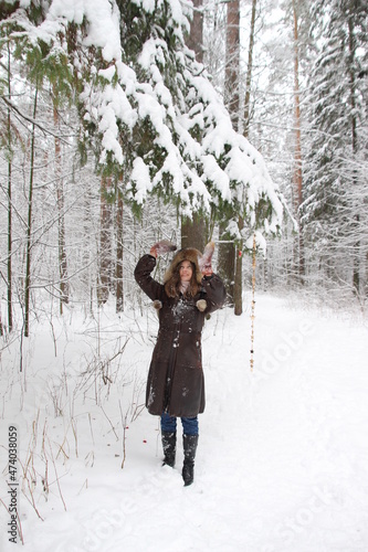 Young pretty woman in brown fur coat, hat, mittens makes snow fall from Chritmas fir tree and smiling in russian winter forest