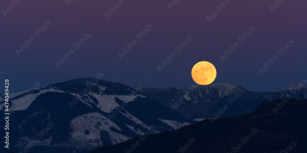 A winter wolf moon touches a mountain ridge with patches of snow where trees have been clearcut. The moon is pink from the recent sunset and the sky is purple A winter wolf moon touches a mountain ridge with patches of snow where trees have been clearcut. The moon is pink from the recent sunset and the sky is purple