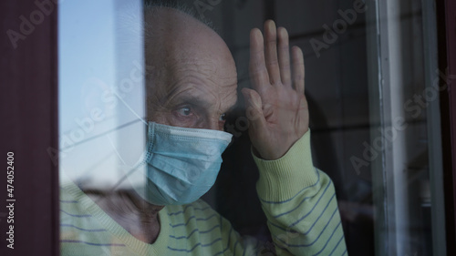 Lonely mature grandfather in facemask looking through closed window