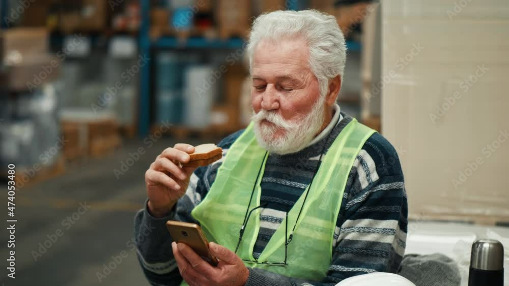 Old man uses a smartphone and eats a sandwich at work during a break Old man uses a smartphone and eats a sandwich at work during a break