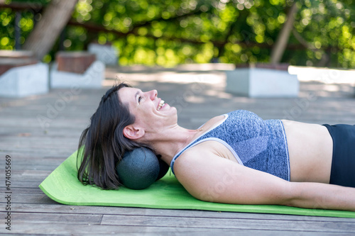 Cheerful athletic girl lies on yoga mat and does neck massage with myofascial balls. Kneads the muscles of neck with MFR roller outdoors photo