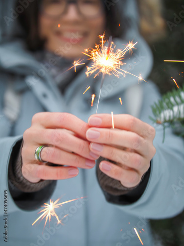 Smiling woman celebrates Christmas or New Year with bright sparkler. Bengal fire, traditional firework for winter holiday celebration outdoors.