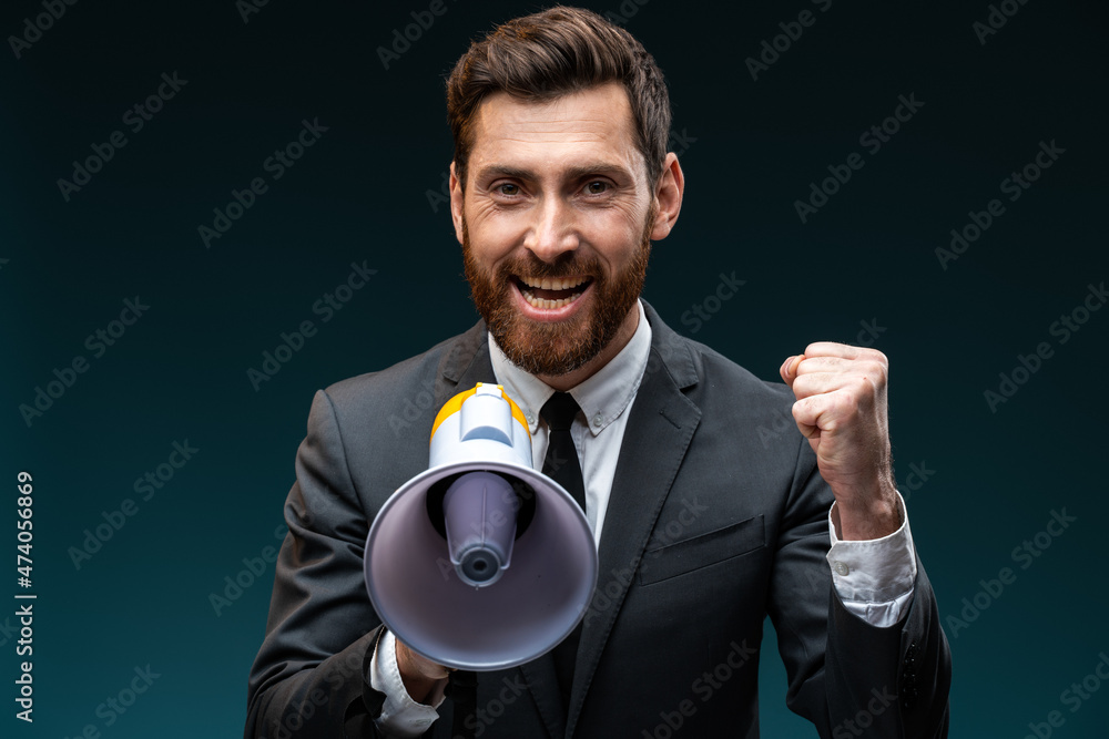 Happy overjoyed man with beard in suit loudly screaming at megaphone, making announce, protesting, wants to be heard. Indoor studio shot isolated on blue background Happy overjoyed man with beard in suit loudly screaming at megaphone, making announce, protesting, wants to be heard. Indoor studio shot isolated on blue background