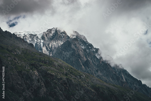 Dark atmospheric surreal landscape with dark rocky mountain top in low clouds in gray cloudy sky. Gray low cloud on high pinnacle. High black rock with snow in low clouds. Surrealist gloomy mountains.