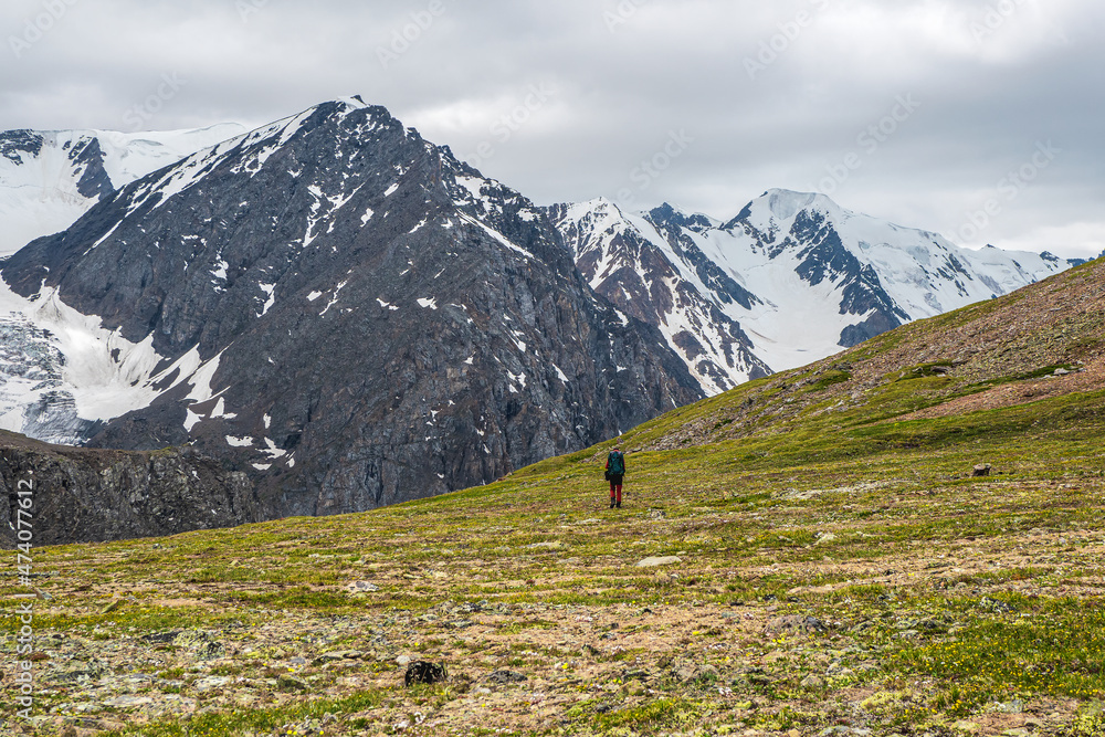 Alpine highlands. Man climbs a mountain slope. In the background is a dome rocky mountains under a cloudy sky.
