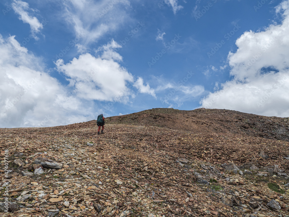 Long solo hike in the highlands. Man climbs a mountain slope. In the background is a dome rocky  mountains under a blue cloudy sky.