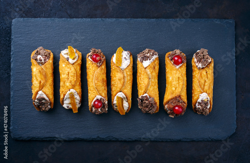 Traditional Italian Sicilian pastry dessert cannoli with creamy ricotta filling, chocolate crisps and fresh currant fruit served as top view on a rustic black board photo