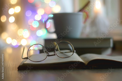 Cup of warm drink with cinnamon stick and candy cane, open book, reading glasses, lit candles and colorful bokeh lights. Selective focus. © jelena990