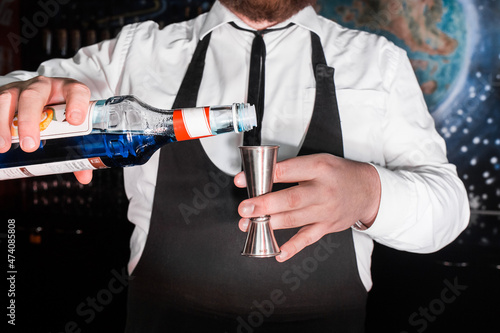 The hand of a professional bartender pours blue syrup into a tool to control the ingredients added to a jigger cocktail or measuring cup. The process of preparing a cocktail